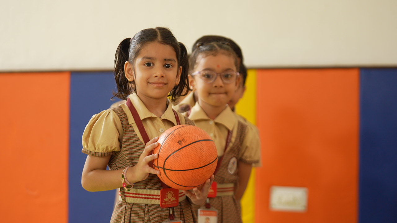 A smiling kindergarten girl holding a basketball during a group activity at SKS World School