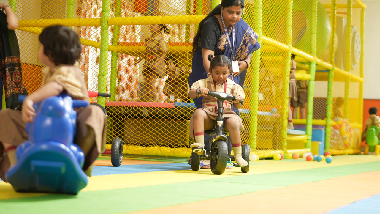 A nursery child riding a tricycle in the play zone while a teacher looks on
